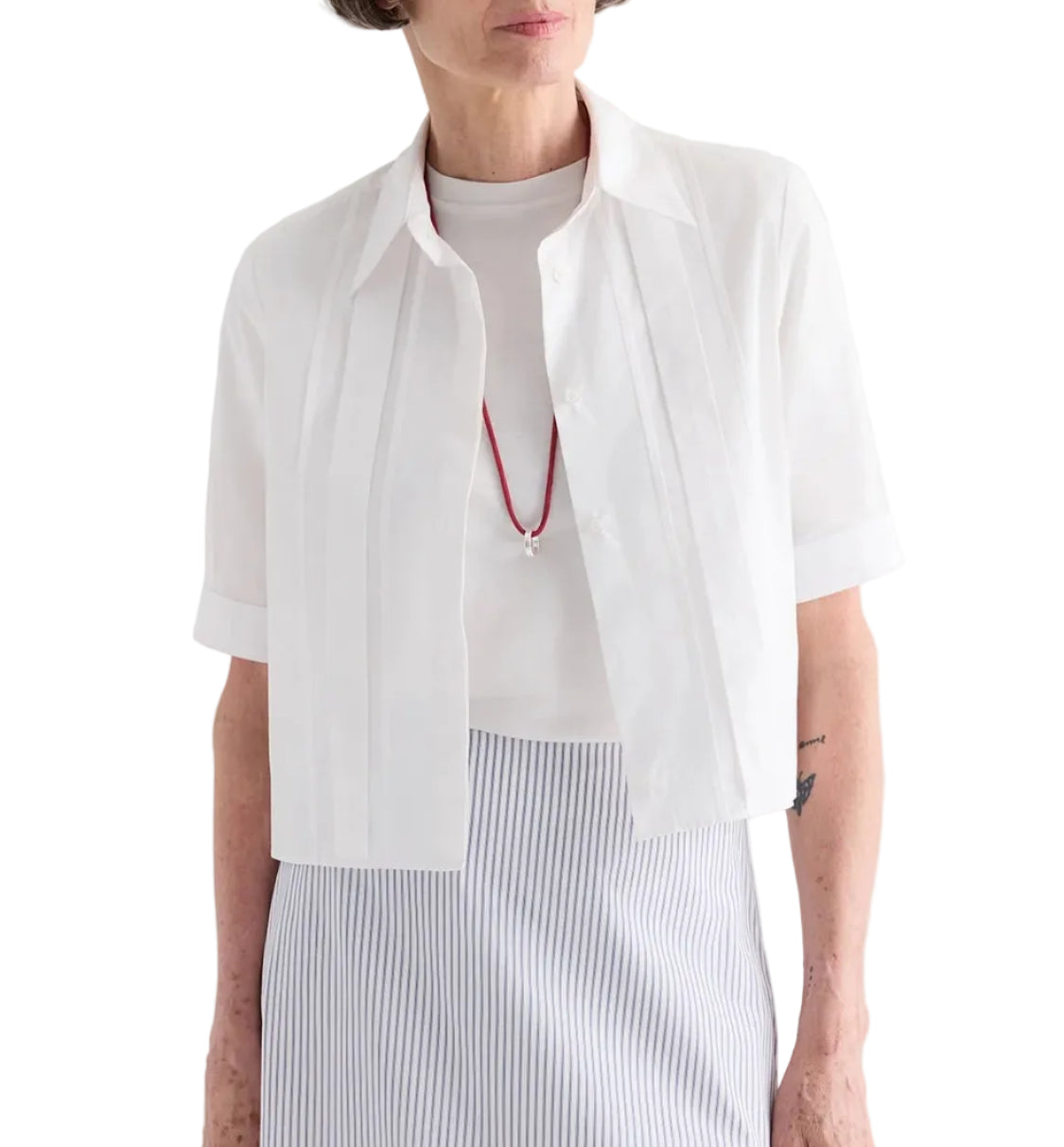 A woman wears the TWP Short Lived Cherry Pleat Top over a white top, paired with a red necklace and a light blue striped skirt, set against a white background.