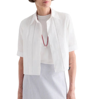 A woman wears the TWP Short Lived Cherry Pleat Top over a white top, paired with a red necklace and a light blue striped skirt, set against a white background.