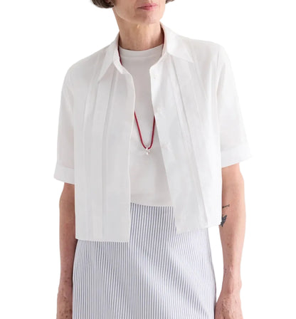 A woman wears the TWP Short Lived Cherry Pleat Top over a white top, paired with a red necklace and a light blue striped skirt, set against a white background.