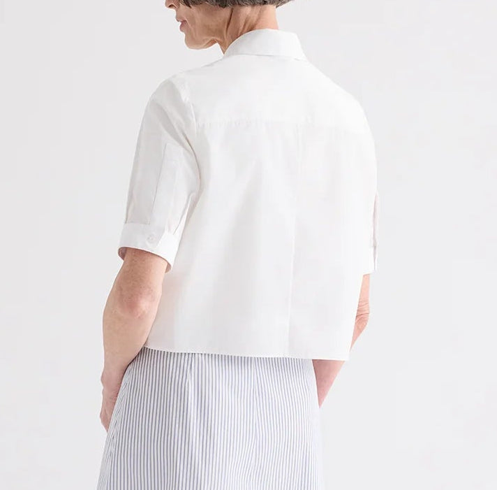 A person shown from the back wears the TWP Short Lived Cherry Pleat Top, a white cropped button-down shirt, paired with a light blue striped skirt against a plain background.