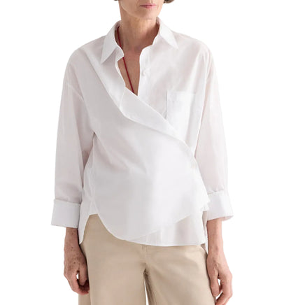 A person wearing the TWP Newest Earl Shirt—a white, wrap-style, long-sleeve shirt crafted from superfine cotton—paired with beige pants, stands against a white background.