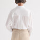 A person with short hair is shown from behind wearing the TWP Newest Earl Shirt, an oversized button-down in white superfine cotton, paired with beige pants and standing against a plain white background.