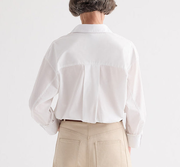 A person with short hair is shown from behind wearing the TWP Newest Earl Shirt, an oversized button-down in white superfine cotton, paired with beige pants and standing against a plain white background.