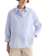 Wearing the TWP Newest Earl Shirt in Mini Banker Stripe—an oversized light blue button-down with long sleeves, a chest pocket, and an asymmetric button front—styled over a white top and beige pants.
