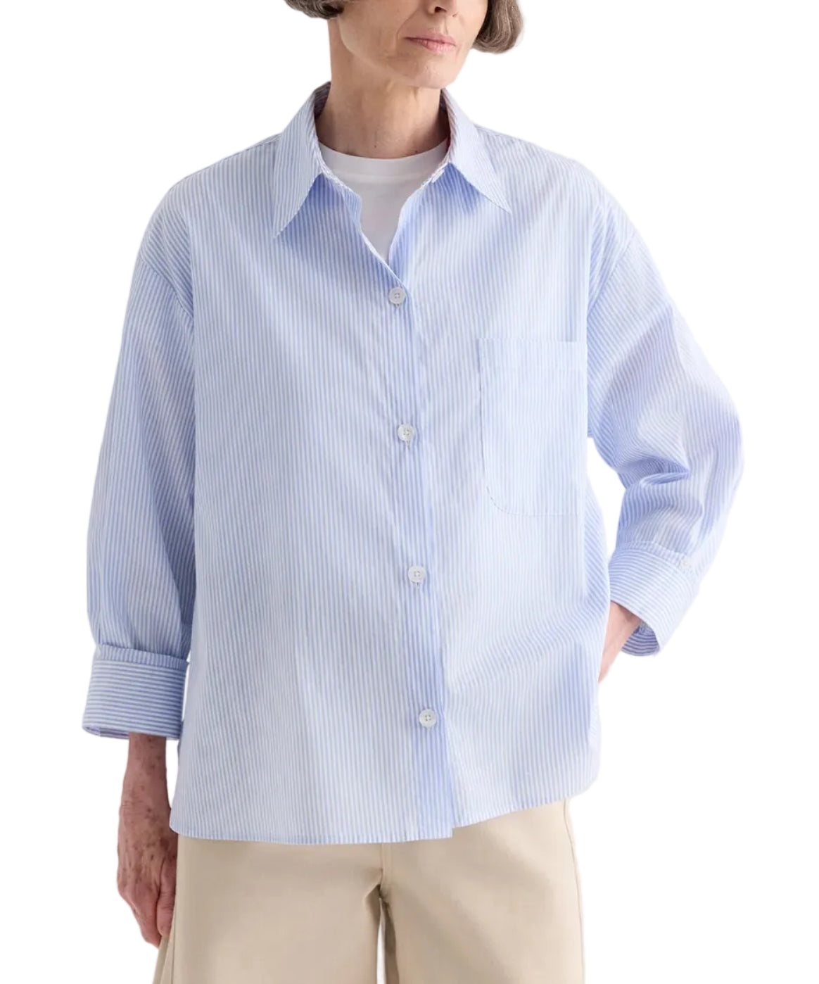 Wearing the TWP Newest Earl Shirt in Mini Banker Stripe—an oversized light blue button-down with long sleeves, a chest pocket, and an asymmetric button front—styled over a white top and beige pants.