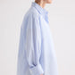 A person stands in profile against a plain background, wearing the TWP Newest Earl Shirt in Mini Banker Stripe—an oversized, light blue and white button-down with the sleeves rolled up.
