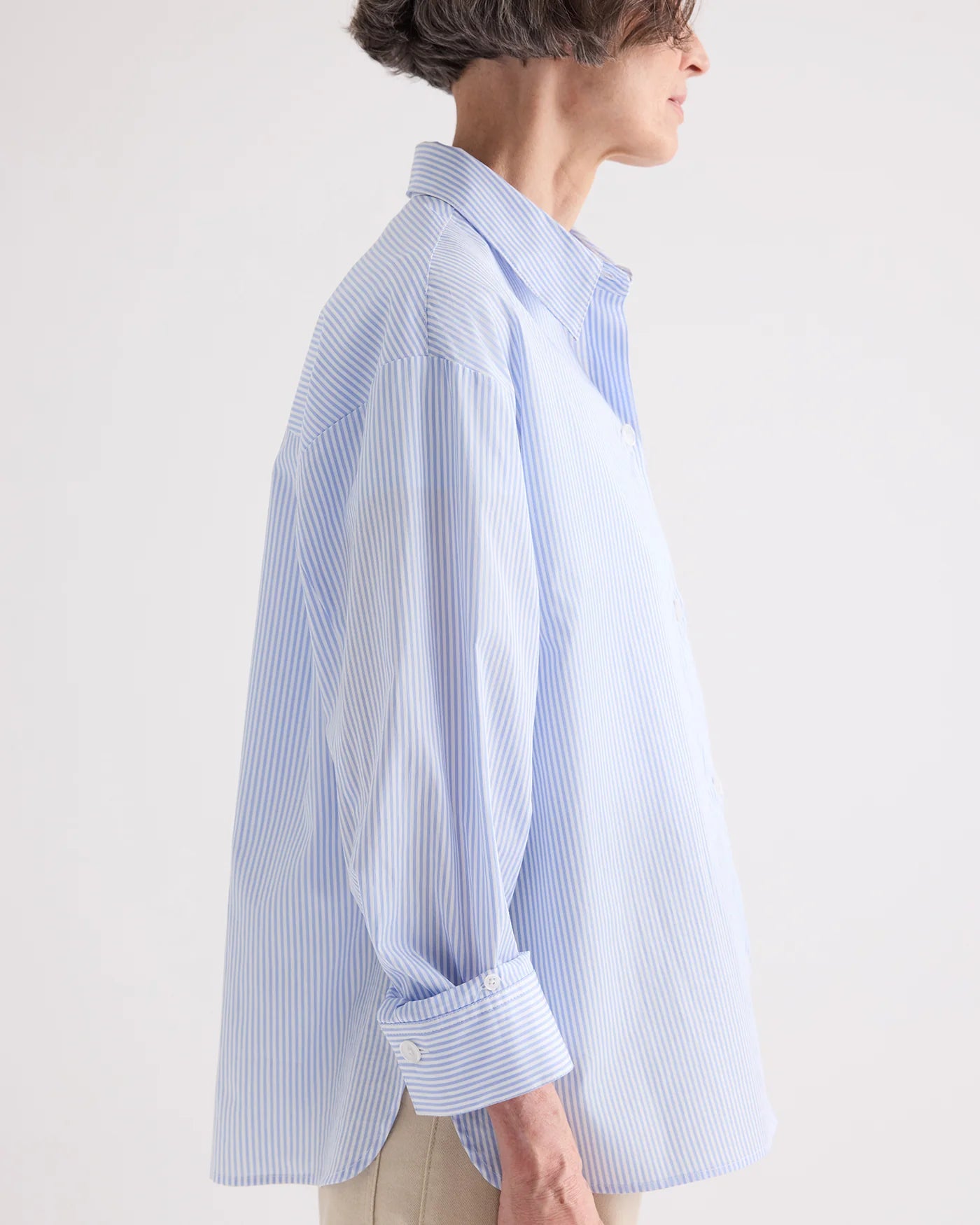 A person stands in profile against a plain background, wearing the TWP Newest Earl Shirt in Mini Banker Stripe—an oversized, light blue and white button-down with the sleeves rolled up.