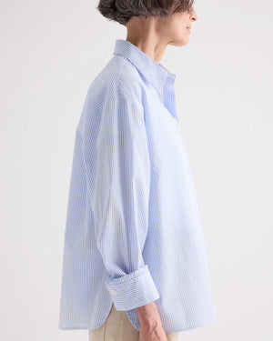 A person stands in profile against a plain background, wearing the TWP Newest Earl Shirt in Mini Banker Stripe—an oversized, light blue and white button-down with the sleeves rolled up.