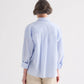 A person with short gray hair, shown from the back against a plain light background, wears TWP’s Newest Earl Shirt in Mini Banker Stripe—an oversized button-down in light blue and white—paired with beige pants.