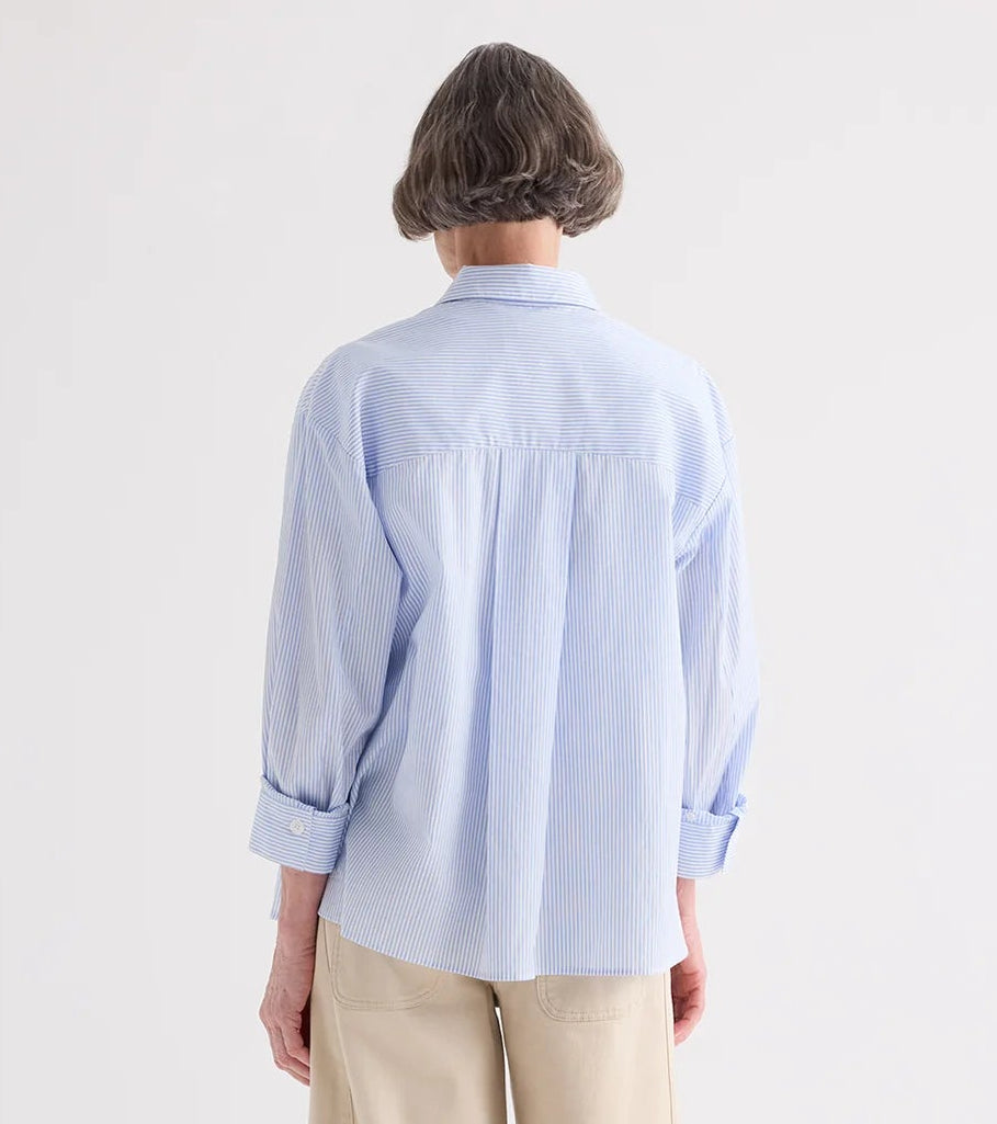 A person with short gray hair, shown from the back against a plain light background, wears TWP’s Newest Earl Shirt in Mini Banker Stripe—an oversized button-down in light blue and white—paired with beige pants.