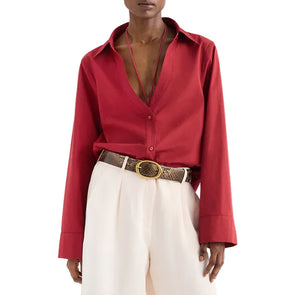 A person wearing a TWP Deep V Relaxed Shirt in Karanda Red, paired with Italian superfine cotton white pants and a brown belt with a large round buckle.