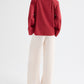 A person stands facing away in the TWP Deep V Relaxed Shirt in Karanda Red Italian superfine cotton, paired with wide-leg cream pants, set against a plain white background.
