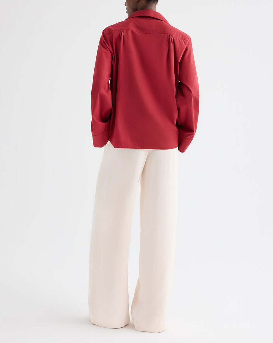 A person stands facing away in the TWP Deep V Relaxed Shirt in Karanda Red Italian superfine cotton, paired with wide-leg cream pants, set against a plain white background.