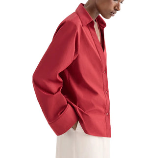 A person wears the Deep V Relaxed Shirt by TWP, crafted from Italian superfine cotton in Karanda Red, paired with off-white pants and standing against a plain background.