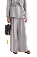 A person wearing a light gray belted jacket paired with the TWP Avery Wool Flannel Pant, holding a black bag with a yellow strap, and wearing brown sandals.
