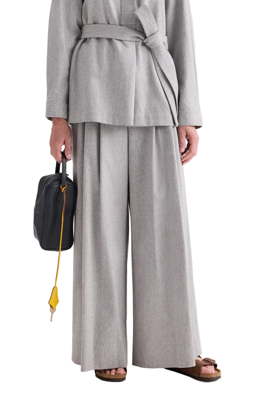 A person wearing a light gray belted jacket paired with the TWP Avery Wool Flannel Pant, holding a black bag with a yellow strap, and wearing brown sandals.
