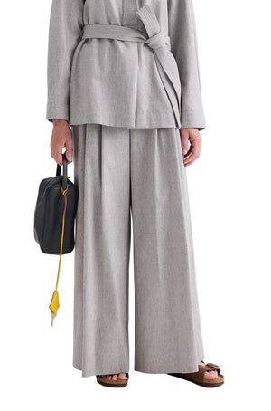 A person wearing a light gray belted jacket paired with the TWP Avery Wool Flannel Pant, holding a black bag with a yellow strap, and wearing brown sandals.