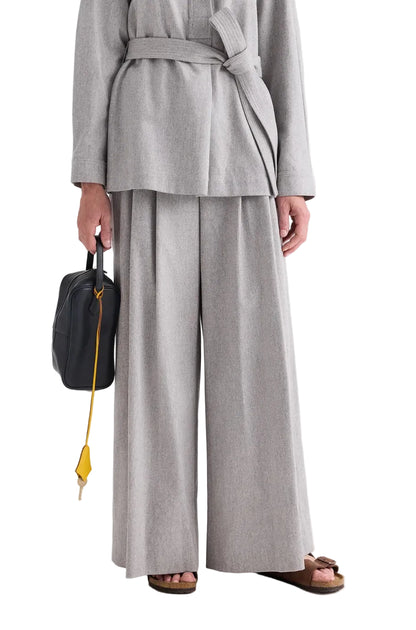 A person wearing a light gray belted jacket paired with the TWP Avery Wool Flannel Pant, holding a black bag with a yellow strap, and wearing brown sandals.