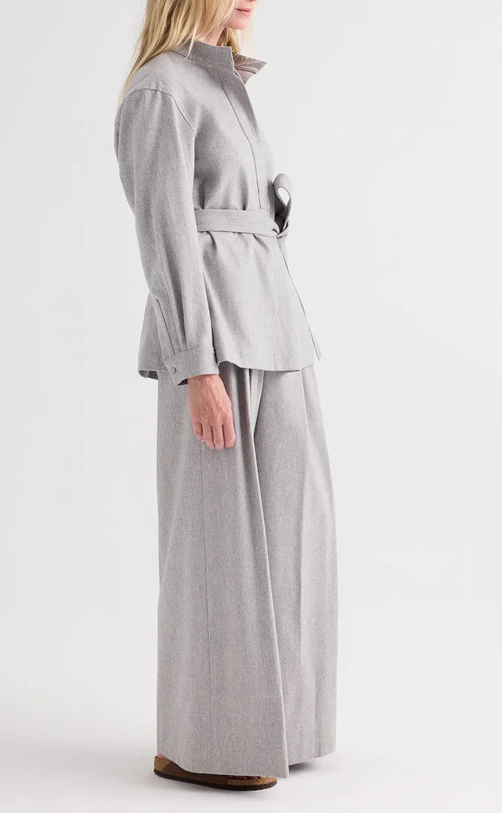 A person stands in profile against a plain white background, wearing TWP’s Avery Wool Flannel Pant—a light gray, mid-rise, wide-leg trouser—paired with a matching belted wool flannel jacket and brown shoes.