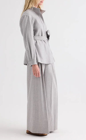 A person stands in profile against a plain white background, wearing TWP’s Avery Wool Flannel Pant—a light gray, mid-rise, wide-leg trouser—paired with a matching belted wool flannel jacket and brown shoes.