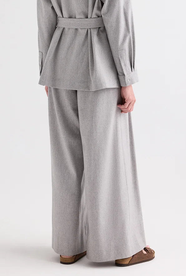 A person is photographed from the back against a plain background, wearing TWP’s Avery Wool Flannel Pant—a light gray, mid-rise, wide-leg style—paired with brown sandals and a loose-fitting suit.