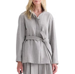 A woman wears the Tamzin Jacket by TWP—a light heather grey, long-sleeve belted wrap jacket crafted from wool flannel—paired with a matching skirt. The image is set against a white background.