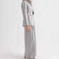 A person stands in profile against a plain white background, wearing the TWP Tamzin Jacket in light heather grey wool flannel with a belted wrap design, paired with matching wide-leg pants and brown sandals.