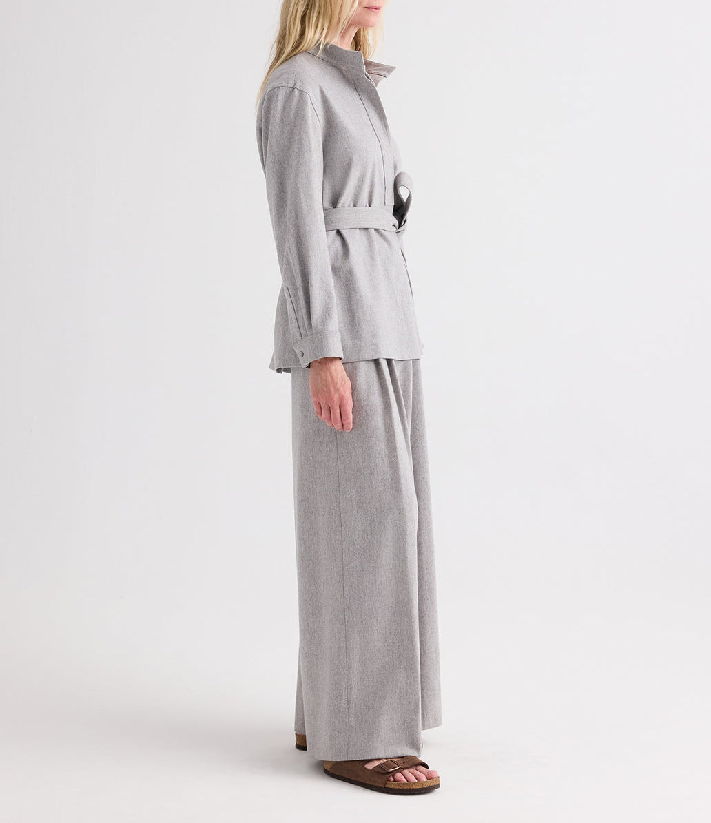 A person stands in profile against a plain white background, wearing the TWP Tamzin Jacket in light heather grey wool flannel with a belted wrap design, paired with matching wide-leg pants and brown sandals.