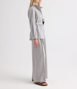 A person stands in profile against a plain white background, wearing the TWP Tamzin Jacket in light heather grey wool flannel with a belted wrap design, paired with matching wide-leg pants and brown sandals.