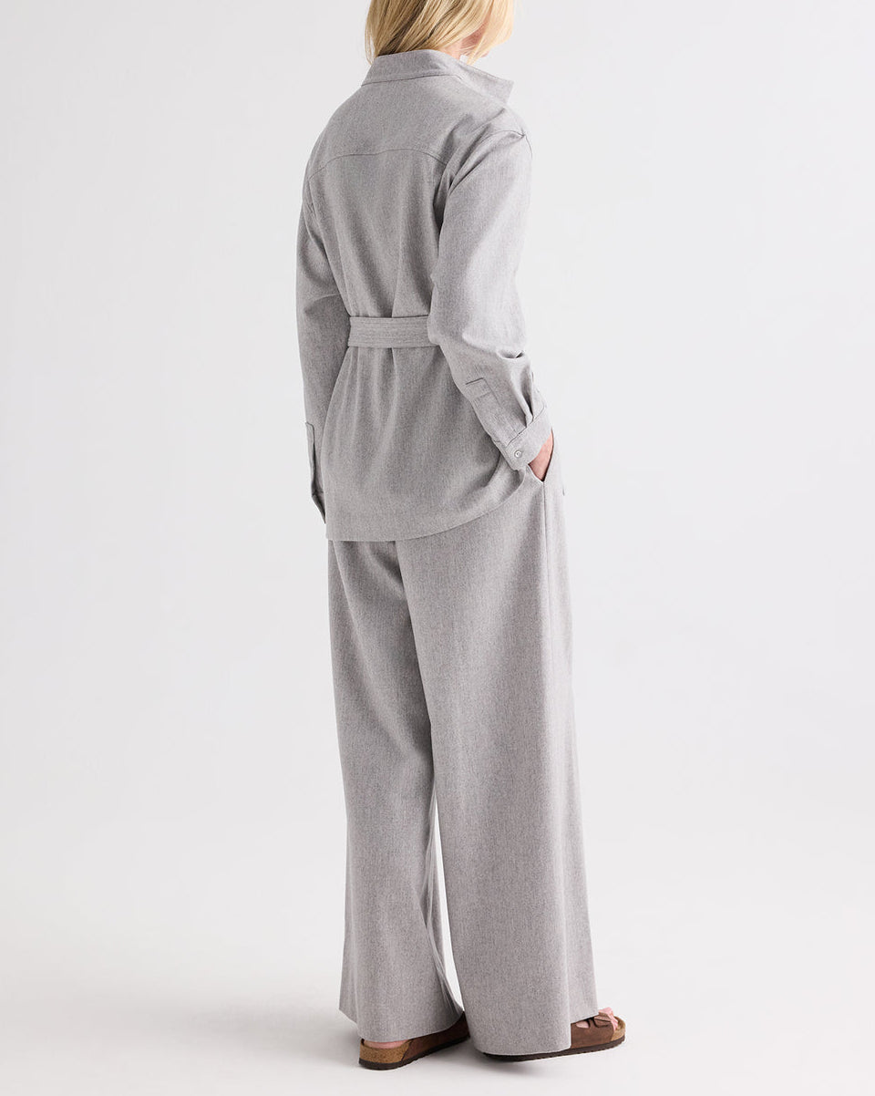A person stands with their back to the camera, wearing the TWP Tamzin Jacket in Light Heather Grey wool flannel, belted at the waist, paired with matching wide-leg pants and brown sandals, set against a plain white background.