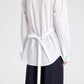 Back view of a white superfine cotton The Night We Met Shirt by TWP with long sleeves and a self-tie waist, worn with dark trousers.