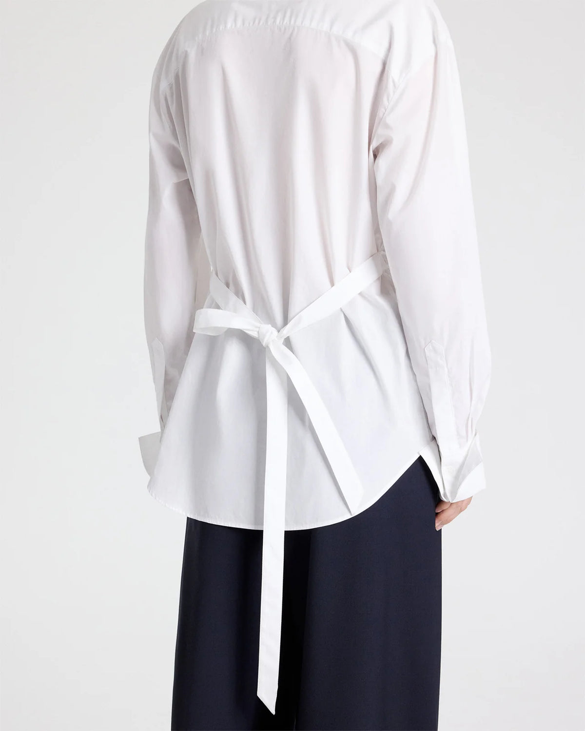 Back view of a white superfine cotton The Night We Met Shirt by TWP with long sleeves and a self-tie waist, worn with dark trousers.