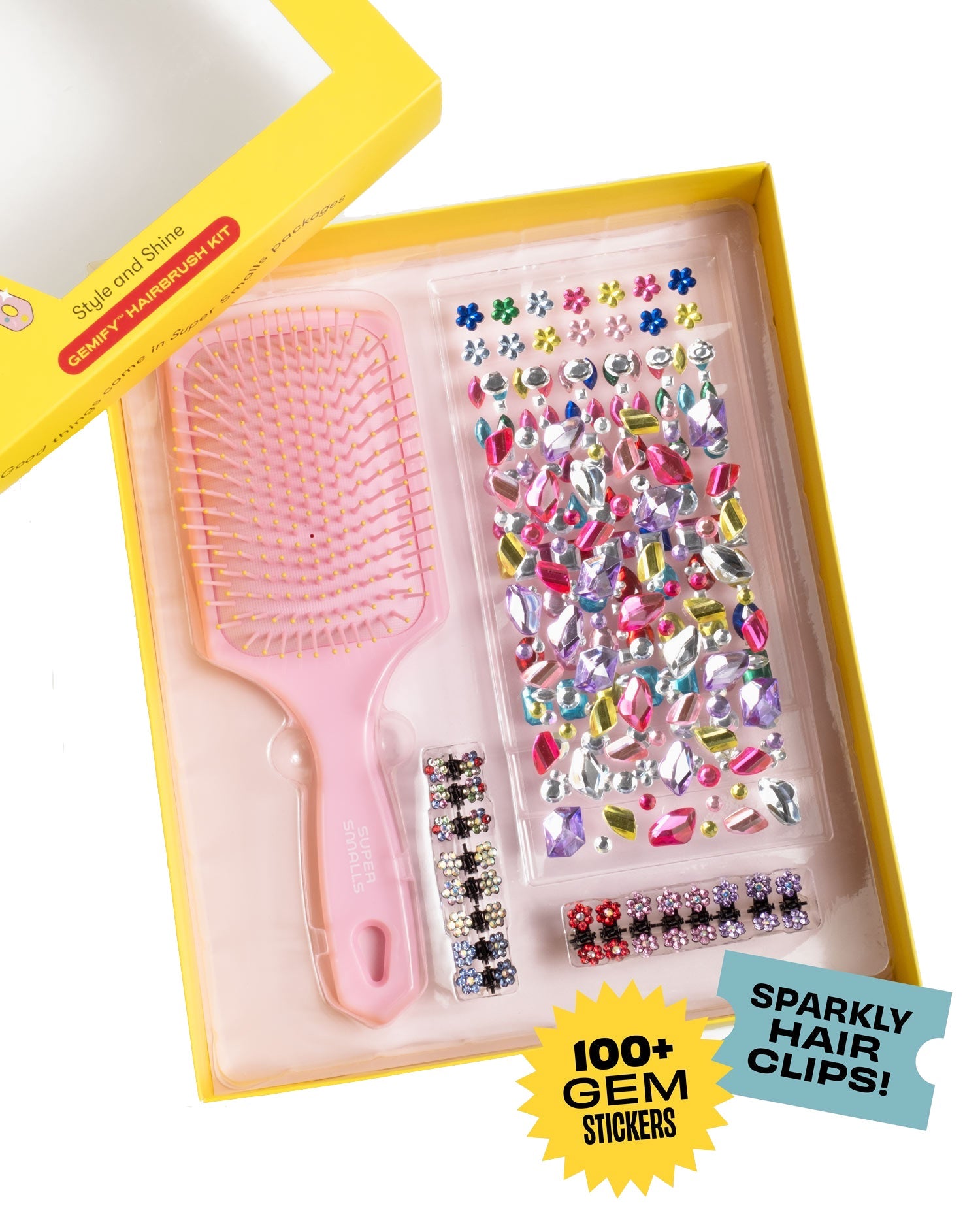 A yellow box from Super Smalls holds a Style and Shine Gemify Hairbrush Kit with a kids hairbrush, colorful gem stickers, and sparkly hair clips. The packaging says "100+ gem stickers" and "Sparkly hair clips!" for creative fun.