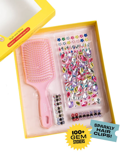 A yellow box from Super Smalls holds a Style and Shine Gemify Hairbrush Kit with a kids hairbrush, colorful gem stickers, and sparkly hair clips. The packaging says "100+ gem stickers" and "Sparkly hair clips!" for creative fun.
