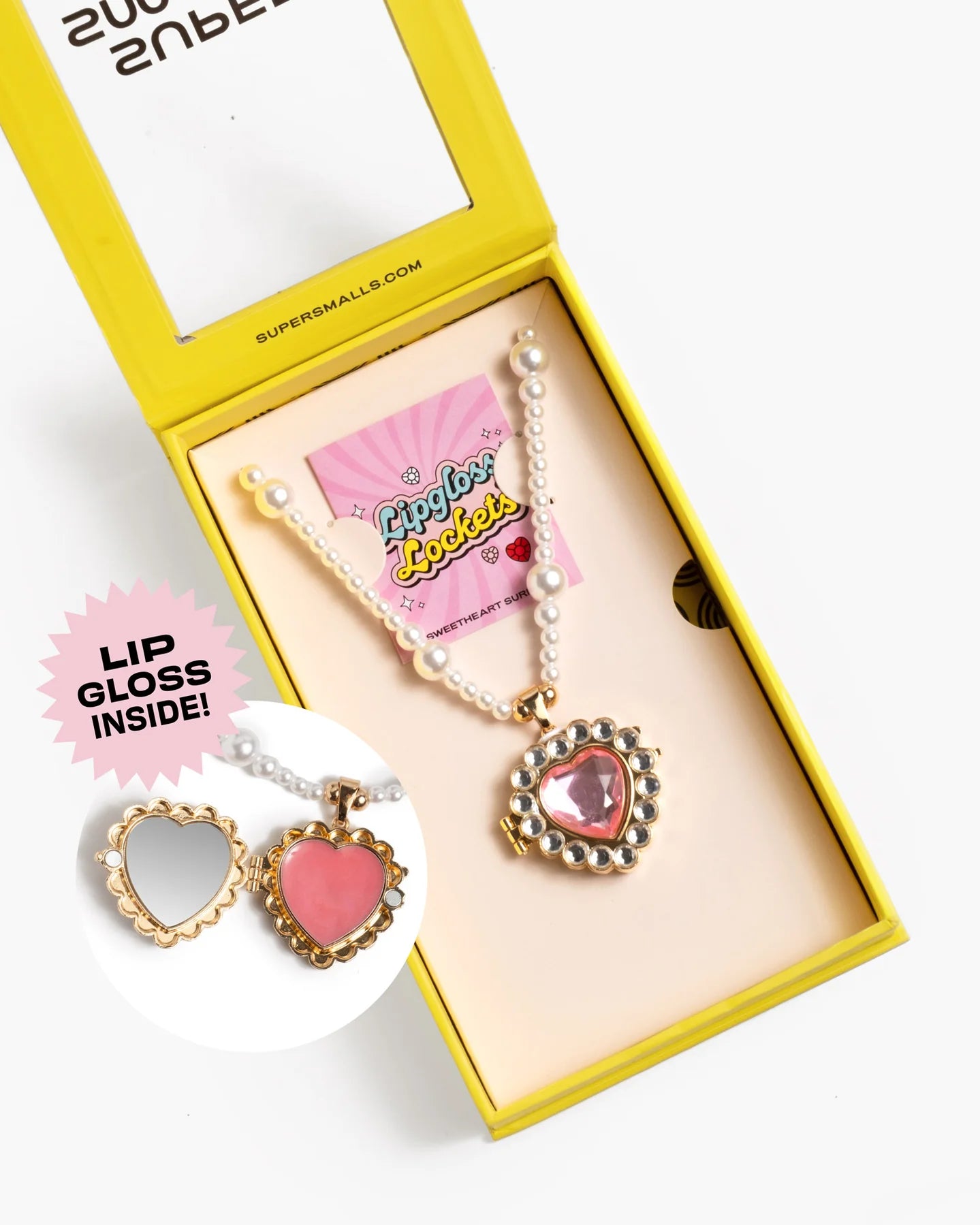 The Sweetheart Surprise Lipgloss Locket by Super Smalls features a heart-shaped compact with pink lip gloss, on a faux-pearl necklace. Presented in a yellow box, it also includes an illustrated card.