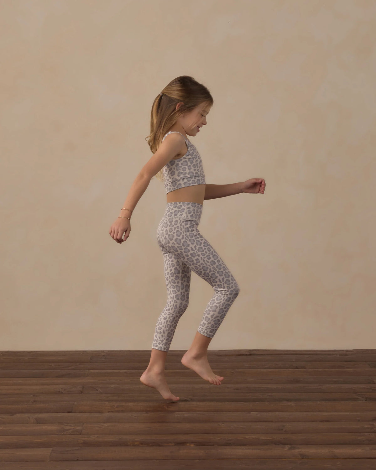 A young girl in Rylee & Cru Girls’ Basic Legging with a leopard print jumps indoors on a wooden floor against a beige background, capturing the energy of everyday flow and effortless class.