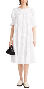 A woman wearing the Merlette Paradis Dress by Merlette New York, with OEKO-TEX® certified cotton lace trim, holding a black handbag, and donning two-toned shoes.