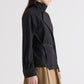 A model is shown from the side against a plain background, wearing The Last Chapter Italian Wool Twill Jacket by TWP—a black belted jacket with a high collar—paired with a beige skirt.