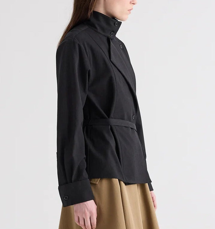 A model is shown from the side against a plain background, wearing The Last Chapter Italian Wool Twill Jacket by TWP—a black belted jacket with a high collar—paired with a beige skirt.
