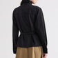 A person is shown from the back wearing the TWP ‘The Last Chapter’ Italian Wool Twill Jacket in black, featuring long sleeves and a belt, paired with a brown skirt, standing against a plain light background.