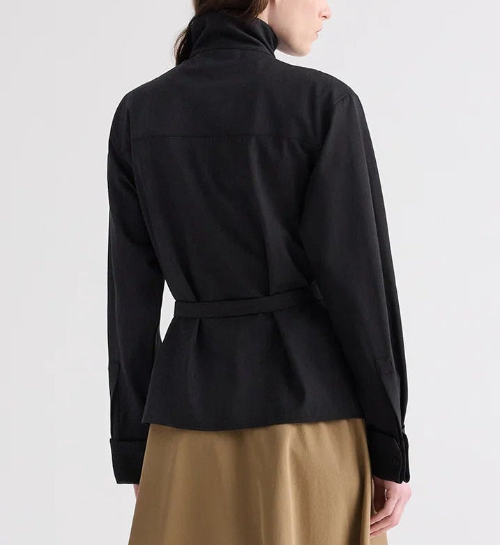 A person is shown from the back wearing the TWP ‘The Last Chapter’ Italian Wool Twill Jacket in black, featuring long sleeves and a belt, paired with a brown skirt, standing against a plain light background.