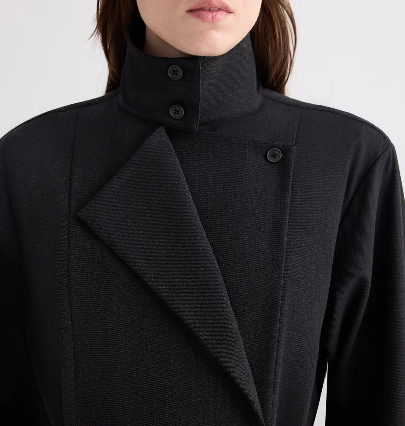 A person is shown from shoulders to mouth wearing TWP’s The Last Chapter Italian Wool Twill Jacket, featuring a high collar, black fabric, an asymmetrical front, and button detailing.