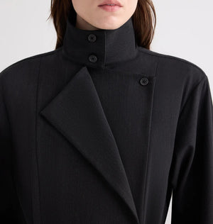 A person is shown from shoulders to mouth wearing TWP’s The Last Chapter Italian Wool Twill Jacket, featuring a high collar, black fabric, an asymmetrical front, and button detailing.