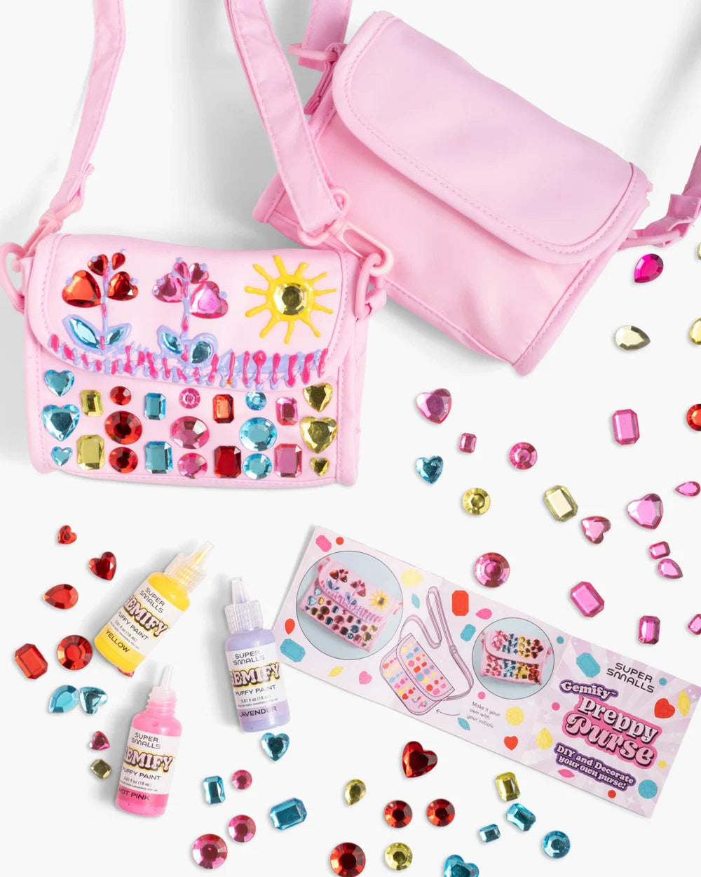 Two pink purses—one adorned with vibrant plastic gems and playful doodles—are surrounded by loose gems, gem glue, and an instruction sheet on a white background, showcasing the Super Smalls Preppy Purse Gemify™ Craft Kit: a delightful DIY gem purse set for creative fun.