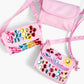 Three small pink shoulder bags from the Super Smalls Preppy Purse Gemify™ Craft Kit—two decorated with colorful gem stickers and drawings, and one plain DIY gem purse—are arranged on a white background.