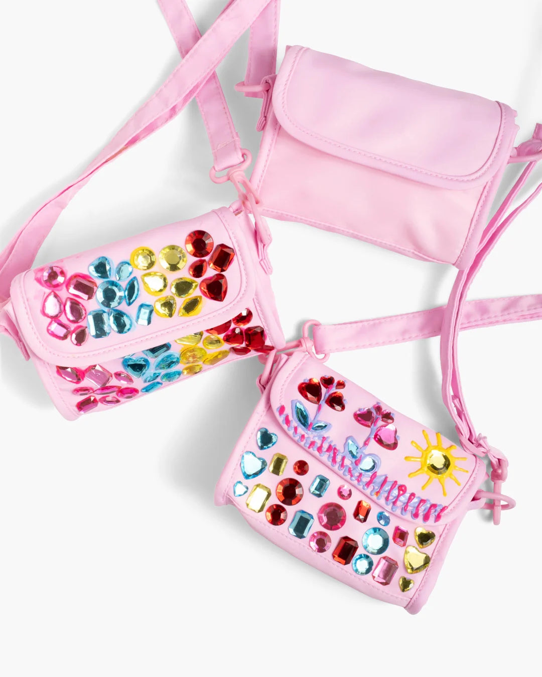Three small pink shoulder bags from the Super Smalls Preppy Purse Gemify™ Craft Kit—two decorated with colorful gem stickers and drawings, and one plain DIY gem purse—are arranged on a white background.