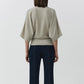 A person stands with their back to the camera, wearing the Simkhai Shai Tee—a light beige cashmere dolman-sleeve top—paired with dark blue jeans, set against a plain white background.