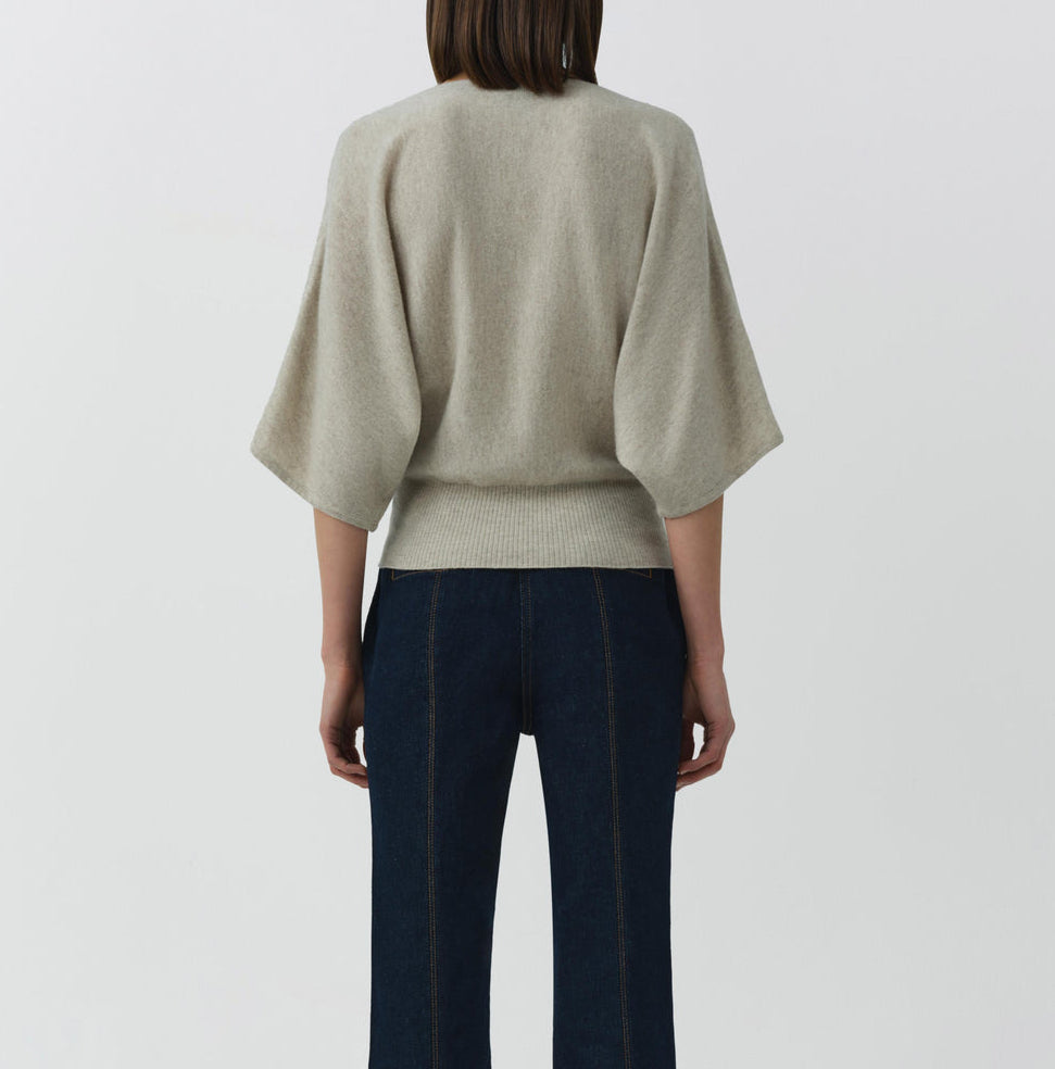 A person stands with their back to the camera, wearing the Simkhai Shai Tee—a light beige cashmere dolman-sleeve top—paired with dark blue jeans, set against a plain white background.