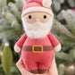 A hand holds the Cuddle + Kind Baby Santa doll—a charming, knitted Santa with a white beard, pink suit, and yellow belt—beside a soft cotton pumpkin baby companion, set against the backdrop of a blurred Christmas tree.