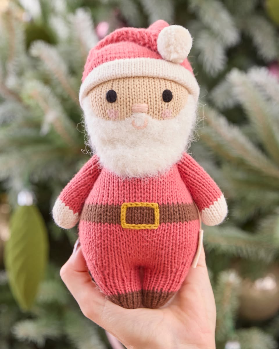 A hand holds the Cuddle + Kind Baby Santa doll—a charming, knitted Santa with a white beard, pink suit, and yellow belt—beside a soft cotton pumpkin baby companion, set against the backdrop of a blurred Christmas tree.
